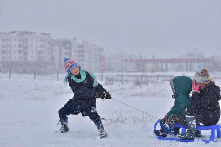 Three children enjoying winter snow, one pulling a sled with buildings in the background.